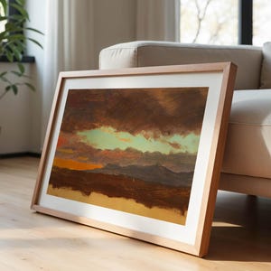 May include: A framed landscape painting with a light wood frame. The artwork depicts a dramatic sky with dark brown clouds, a band of green, and an orange sunset over a mountain range. The painting leans against a beige sofa.