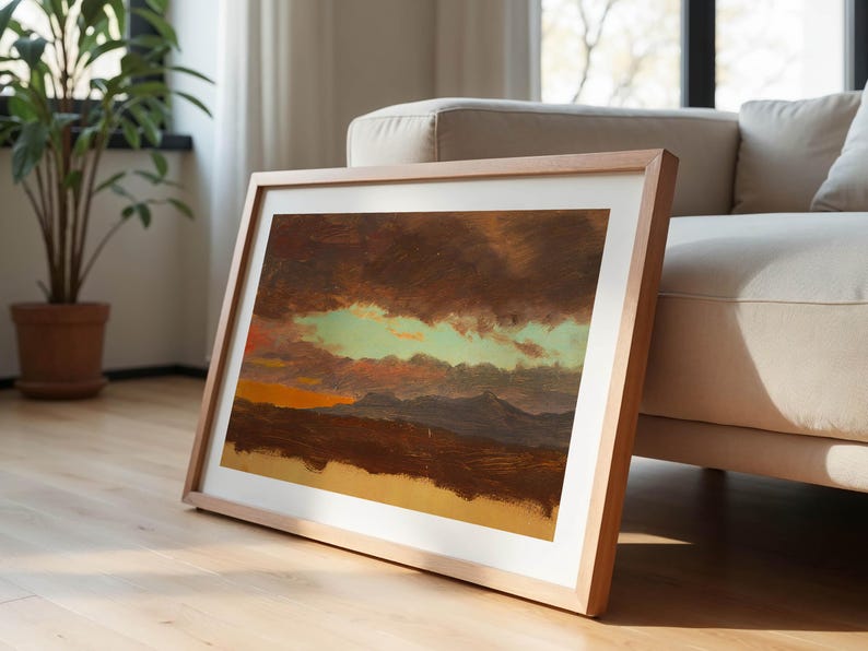 May include: A framed landscape painting with a light wood frame. The artwork depicts a dramatic sky with dark brown clouds, a band of green, and an orange sunset over a mountain range. The painting leans against a beige sofa.
