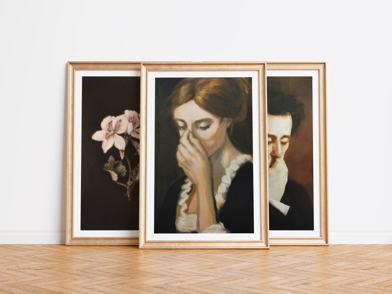 Op de afbeelding: Drie ingelijste kunstprints met gouden lijsten. De linker print toont een roze bloem op een donkere achtergrond. De middelste print toont een vrouw met rood haar die naar beneden kijkt, haar hand bedekt haar mond. De rechter print toont een man met donker haar die naar beneden kijkt, zijn hand bedekt zijn mond.