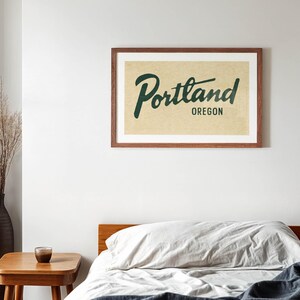 May include: A framed print with the word "Portland" in a cursive font and "Oregon" below it, in a dark green color. The print is in a brown wooden frame and hangs on a white wall above a bed with a wooden frame and a gray comforter.