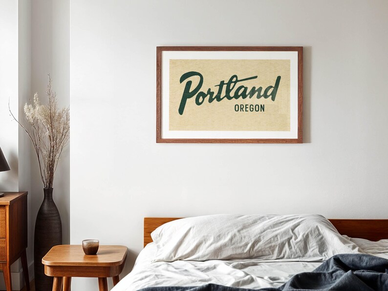 May include: A framed print with the word "Portland" in a cursive font and "Oregon" below it, in a dark green color. The print is in a brown wooden frame and hangs on a white wall above a bed with a wooden frame and a gray comforter.