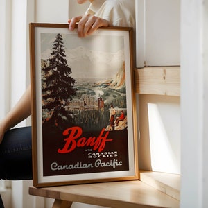 May include: A framed vintage travel poster advertising Banff in the Canadian Rockies. The poster features a mountain scene with a large evergreen tree in the foreground, a river, and a hotel in the distance. The text on the poster reads "Banff in the Canadian Rockies" and "Canadian Pacific".