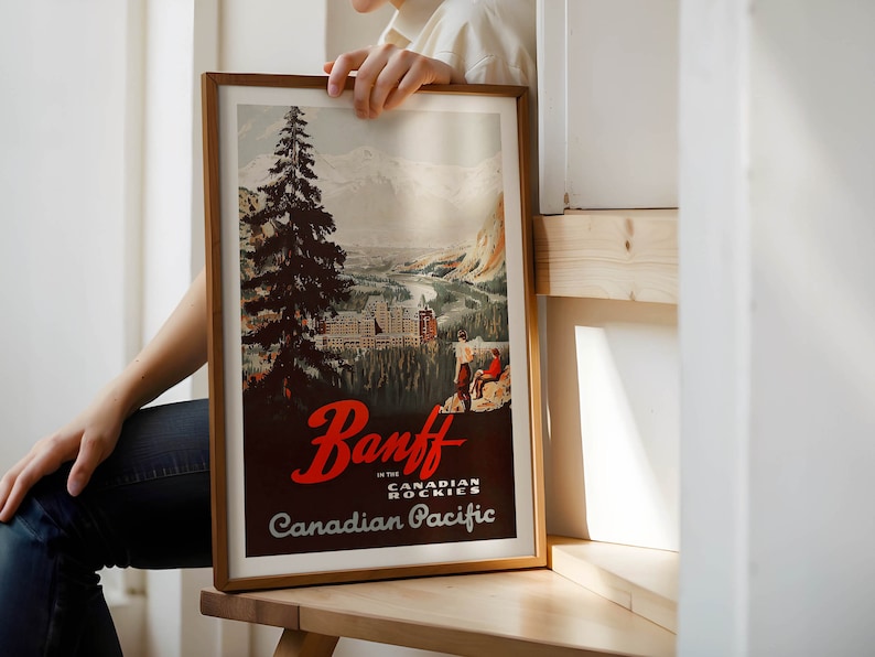 May include: A framed vintage travel poster advertising Banff in the Canadian Rockies. The poster features a mountain scene with a large evergreen tree in the foreground, a river, and a hotel in the distance. The text on the poster reads "Banff in the Canadian Rockies" and "Canadian Pacific".