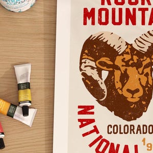 Pu&ograve; includere: Un poster in stile vintage con un ariete marrone e beige con grandi corna. Il testo "Rocky Mountain National Park Colorado 1991" &egrave; stampato in rosso e bianco.