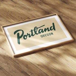 May include: A framed print featuring the word "Portland" in a stylized green font, with "Oregon" below it. The print has a cream-colored background and a wooden frame. The artwork is displayed on a wooden surface.