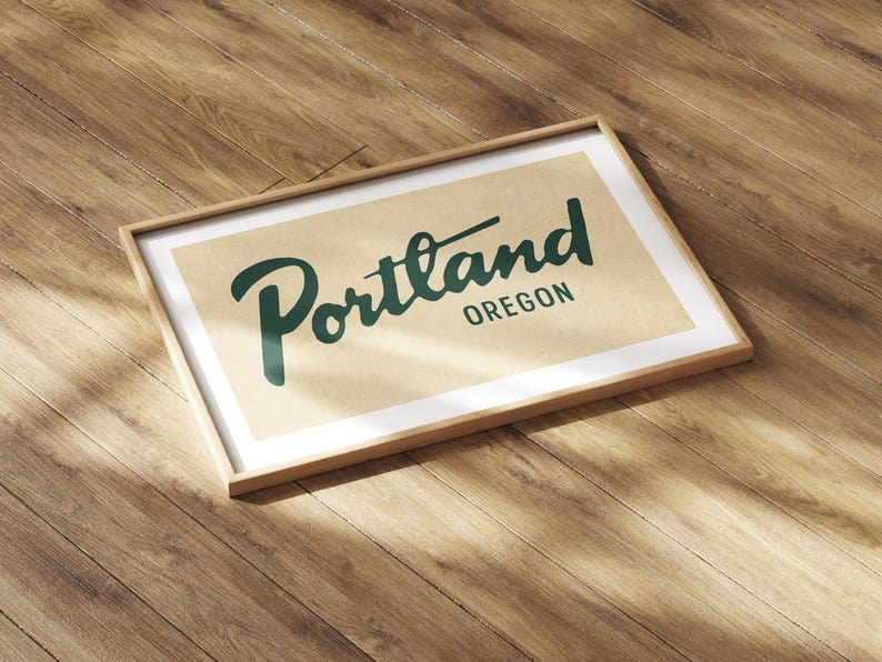 May include: A framed print featuring the word "Portland" in a stylized green font, with "Oregon" below it. The print has a cream-colored background and a wooden frame. The artwork is displayed on a wooden surface.