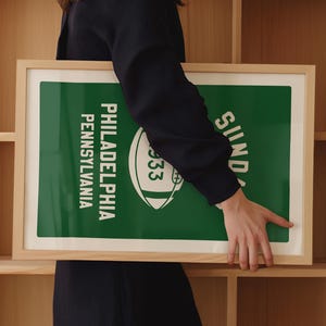 K&ouml;nnte beinhalten: Ein gerahmtes gr&uuml;n-wei&szlig;es Poster mit dem Text "Philadelphia, Pennsylvania" und "Sonntag" sowie einer Fu&szlig;ballgrafik. Das Poster wird von einer Person gehalten.