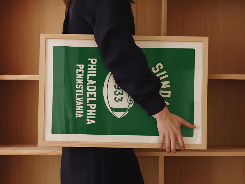 K&ouml;nnte beinhalten: Ein gerahmtes gr&uuml;n-wei&szlig;es Poster mit dem Text "Philadelphia, Pennsylvania" und "Sonntag" sowie einer Fu&szlig;ballgrafik. Das Poster wird von einer Person gehalten.