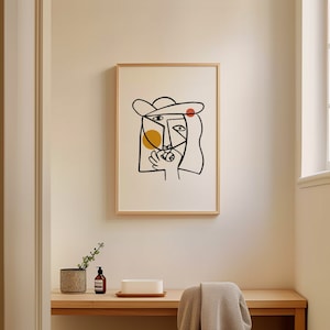 May include: A framed black and white line drawing of a face with a yellow and red circle inside. The drawing is on a white background and is framed in a light wood frame.