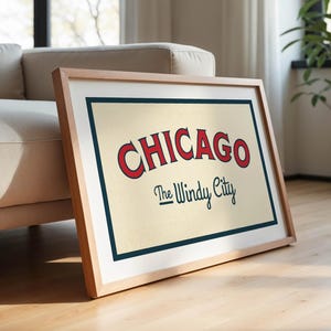 Op de afbeelding: Een ingelijste print met het woord "CHICAGO" in rood, met "The Windy City" eronder in een scriptlettertype. Het kunstwerk heeft een cr&egrave;mekleurige achtergrond met een marineblauwe rand, in een licht houten frame.