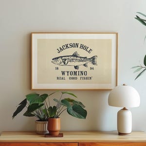 May include: A framed print with a beige background features a detailed illustration of a fish, with the text "JACKSON HOLE WYOMING REAL GOOD FISHIN'". The artwork is in a wooden frame, displayed above a wooden table.