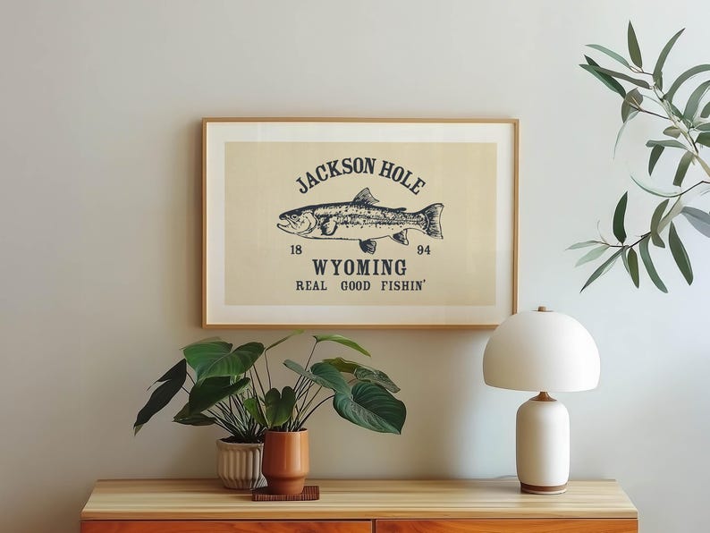 May include: A framed print with a beige background features a detailed illustration of a fish, with the text "JACKSON HOLE WYOMING REAL GOOD FISHIN'". The artwork is in a wooden frame, displayed above a wooden table.