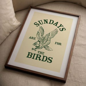 May include: Framed art print with a cream background and a green eagle illustration. The text "SUNDAYS ARE FOR THE BIRDS" is printed above and below the eagle. The frame is dark brown.