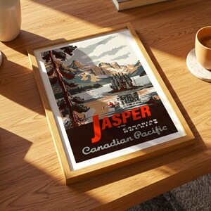 Op de afbeelding: Een ingelijste kunstafdruk met een vintage reisaffiche voor Jasper in de Canadese Rockies. De poster toont een landschap met bergen, een meer en de tekst "Jasper in the Canadian Pacific". De lijst is van licht hout.