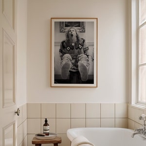 Op de afbeelding: Een zwart-witfoto in een lijst van een persoon die met uitgestrekte benen zit. De persoon draagt een trui met patroon en mocassins. De foto hangt aan een muur in een badkamer.