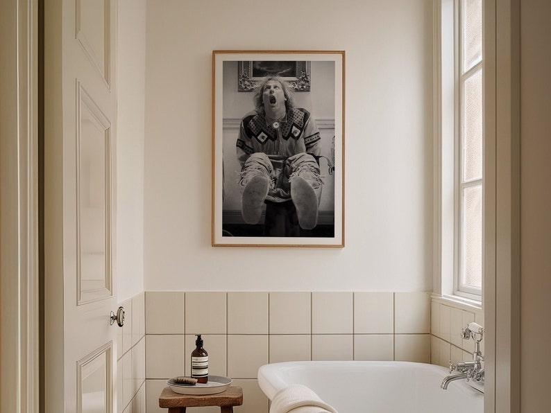 Op de afbeelding: Een zwart-witfoto in een lijst van een persoon die met uitgestrekte benen zit. De persoon draagt een trui met patroon en mocassins. De foto hangt aan een muur in een badkamer.