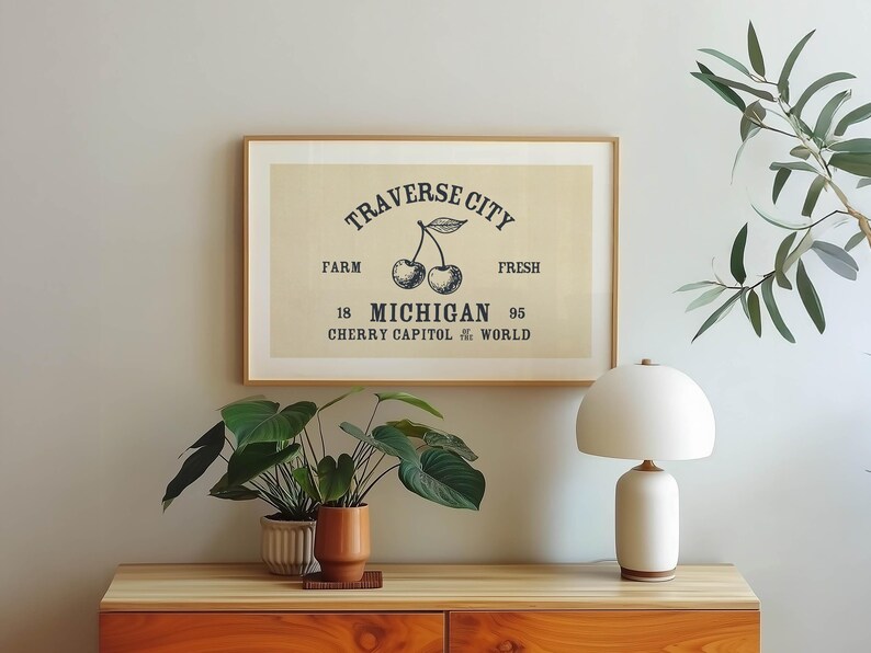 May include: A framed print with the text "Traverse City" and a cherry illustration. The print is in a light-colored frame and hangs above a wooden cabinet. A white lamp and potted plants are also visible.