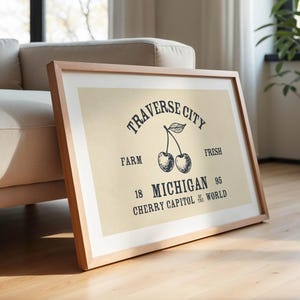 May include: Framed print with a light beige background and a wooden frame. The print features the words "Traverse City" above a cherry illustration. Below the cherries are the words "Farm", "Michigan", and "Cherry Capitol of the World".