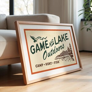 Op de afbeelding: Een ingelijst drukwerk met een vintage ontwerp met de tekst "Game and Lake Outdoors" in groene tekst met een bruine en oranje rand. De print bevat ook een sc&egrave;ne met een meer met bomen en vogels.