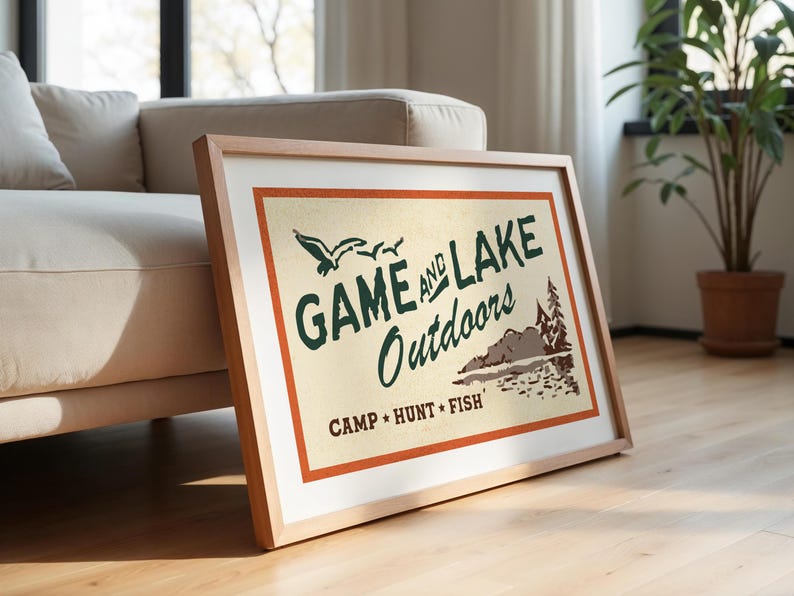 Op de afbeelding: Een ingelijst drukwerk met een vintage ontwerp met de tekst "Game and Lake Outdoors" in groene tekst met een bruine en oranje rand. De print bevat ook een sc&egrave;ne met een meer met bomen en vogels.