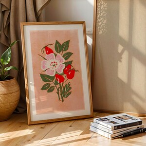 May include: A framed print of a pink and white rose with red berries on a light pink background. The frame is made of wood and has a natural finish.