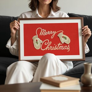 May include: A framed print with a red background and the words "Merry Christmas" in cursive script. Two cream-colored mittens with holly accents are featured. The frame is a dark brown color.