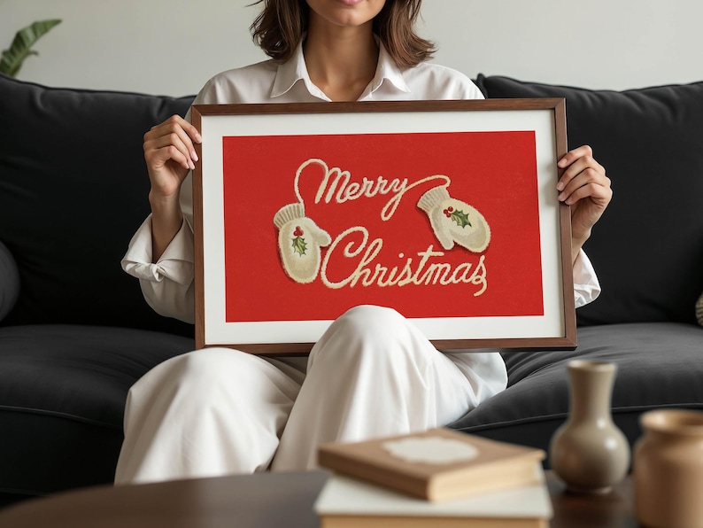 May include: A framed print with a red background and the words "Merry Christmas" in cursive script. Two cream-colored mittens with holly accents are featured. The frame is a dark brown color.