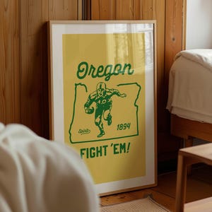 K&ouml;nnte beinhalten: Ein gerahmtes Poster mit gelbem Hintergrund und gr&uuml;nem Text und Grafiken. Das Poster zeigt "Oregon" &uuml;ber einer Footballspieler-Grafik und dem Text "Fight 'Em!". Das Jahr "1894" wird ebenfalls angezeigt.