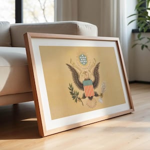K&ouml;nnte beinhalten: Gerahmter Druck des Gro&szlig;en Siegels der Vereinigten Staaten, mit einem Adler mit ausgebreiteten Fl&uuml;geln, Olivenzweig und Pfeilen. Das Kunstwerk wird in einem hellen Holzrahmen pr&auml;sentiert, der an einem beigen Sofa auf einem Parkettboden lehnt.