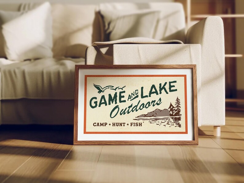 Op de afbeelding: Een ingelijste vintage poster met de tekst "Game and Lake Outdoors" in groene en oranje letters. De poster toont een sc&egrave;ne met een meer, bomen en vogels die erboven vliegen. De tekst "Camp - Hunt - Fish" staat onder de hoofdtitel.