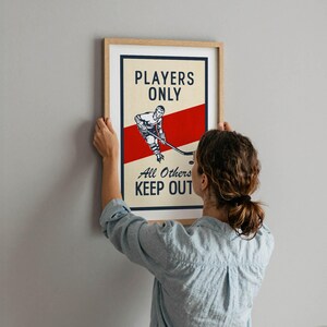 Pu&ograve; includere: Stampa incorniciata a tema hockey con un design vintage. L'opera presenta un'illustrazione di un giocatore di hockey e il testo "PLAYERS ONLY, All Others KEEP OUT" in grassetto. La stampa ha una striscia rossa ed &egrave; tenuta da una persona.
