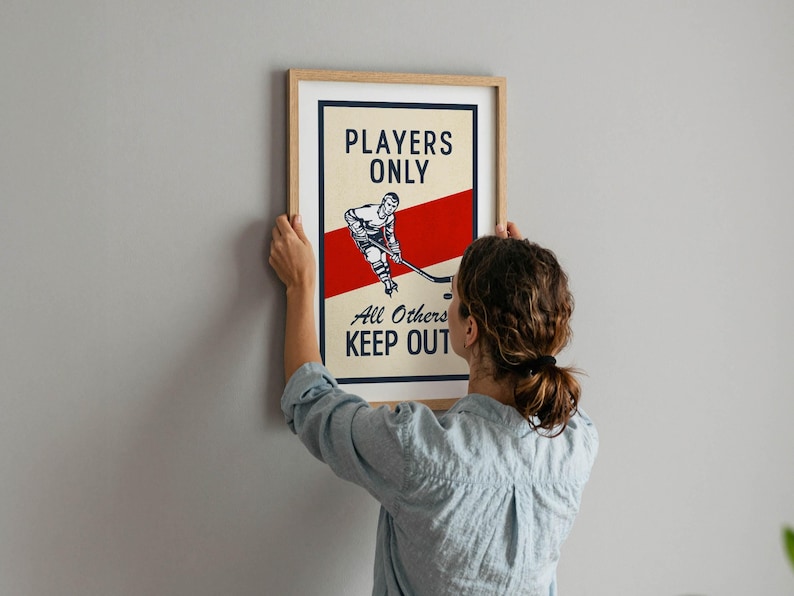 Pu&ograve; includere: Stampa incorniciata a tema hockey con un design vintage. L'opera presenta un'illustrazione di un giocatore di hockey e il testo "PLAYERS ONLY, All Others KEEP OUT" in grassetto. La stampa ha una striscia rossa ed &egrave; tenuta da una persona.
