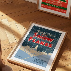 May include: A vintage-style poster with a blue background and white snowflakes. The poster features the text "Santa Claus's Snowflakes" in red and white lettering. The poster is framed in a light wood frame.