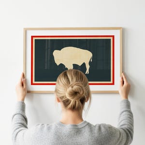 May include: A framed wall art print featuring a light beige silhouette of a bison centered on a dark charcoal background. The artwork is enclosed by a thin red border and a wider cream-colored border, all encased in a light-toned wooden frame.