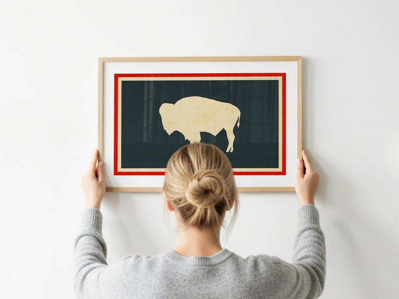 May include: A framed wall art print featuring a light beige silhouette of a bison centered on a dark charcoal background. The artwork is enclosed by a thin red border and a wider cream-colored border, all encased in a light-toned wooden frame.