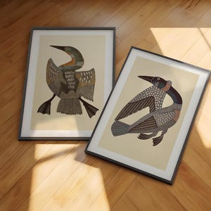 May include: Two framed art prints featuring stylized illustrations of loons. Each print displays a bird with intricate patterns on its wings and body, set against a light cream background. The frames are thin and black, showcasing the detailed avian artwork.