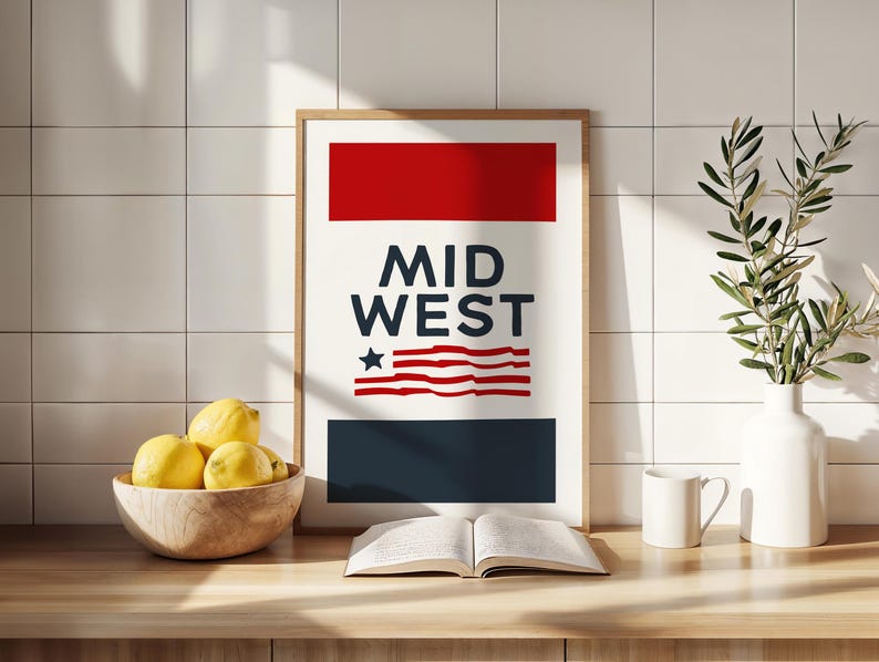 May include: A framed print with a red, white, and blue design. The print says "MID WEST" and features a stylized American flag. The print is on a wooden countertop with a bowl of lemons, a book, a white mug, and a vase with a green plant.