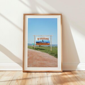 May include: A framed photograph of a sign welcoming visitors to Wyoming. The sign is blue and white with the words "Welcome to Wyoming" and "Forever West" in orange. The sign is on a wooden post and is located on a dirt road.