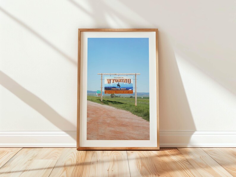 May include: A framed photograph of a sign welcoming visitors to Wyoming. The sign is blue and white with the words "Welcome to Wyoming" and "Forever West" in orange. The sign is on a wooden post and is located on a dirt road.