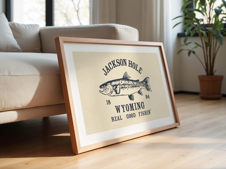 May include: A framed print featuring a vintage-style illustration of a fish, with the text "JACKSON HOLE WYOMING REAL GOOD FISHIN'" in a dark blue color. The print is in a light wood frame and leans against a beige sofa.