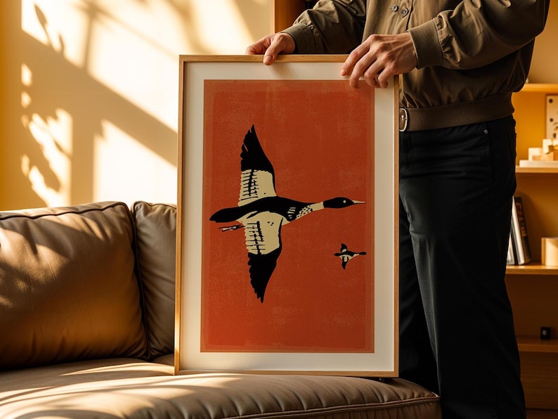 May include: A framed print of a black and white illustration of a bird flying against a red background. The bird has a long neck and a long beak. There is a smaller bird flying below the larger bird.