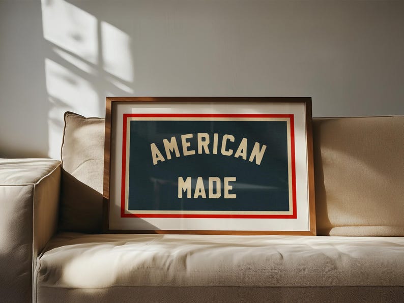 May include: A framed print with the words "AMERICAN MADE" in white on a dark blue background, with a red border. The frame is brown. The print is on a beige sofa, with sunlight creating shadows on the wall.