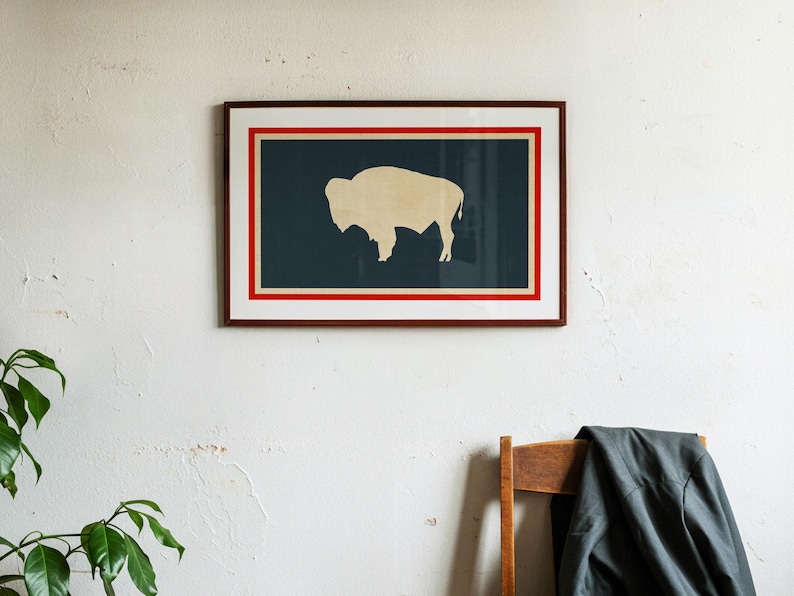 May include: A framed rectangular wall art piece featuring a dark blue background with a cream-colored silhouette of a bison in the center. The artwork is enclosed by a thin red border and a white mat, all contained within a dark brown wooden frame.