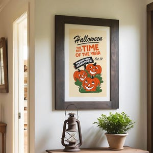 May include: A framed Halloween-themed poster with a dark wood frame. The poster features three smiling orange pumpkins and the text "Halloween, the best time of the year." A vintage lantern and potted plant are on a wooden table.