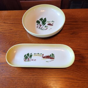 May include: A set of two vintage ceramic dishes with a farmhouse scene. The dishes are white with a yellow rim and feature a green and brown illustration of a farmhouse with trees and a fence.