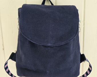 May include: A dark navy blue backpack with a large front flap closure. The bag features adjustable shoulder straps with a repeating pattern of thin horizontal stripes in light purple and dark navy blue, attached to the sides with silver hardware.