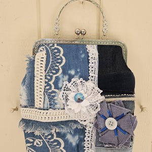 Könnte beinhalten: Handgefertigte Jeans-Geldbörse mit silberfarbenem Metallrahmen und Griff. Die Geldbörse hat ein Patchwork-Design mit weißer Spitze, Häkelbesatz und Stoffblumen. Ein weißer Knopf befindet sich in der Mitte der blauen Stoffblume.