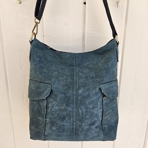 May include: A blue-green shoulder bag with two front pockets. The bag has a dark blue strap and gold-colored metal hardware. The bag is hanging against a white wooden background.