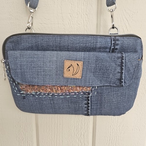 May include: A denim crossbody bag with a front flap and a zipper closure. The bag features a brown patch with a logo and a decorative stitched design. The bag has a denim strap and silver-tone hardware.