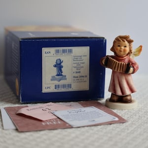 Op de afbeelding: Een porseleinen beeldje van een engel die een rode accordeon bespeelt. De engel draagt een roze jurk en heeft een gele halo. Het beeldje staat op een witte voet en wordt geleverd in een blauwe doos met de tekst "Jahresengel 2000 Engel mit Bandoneon Annual Angel 2000 *Millennium Blue* # 1640 Hum 2096/H 02-096-35-2 12.5 cm/5 inch"